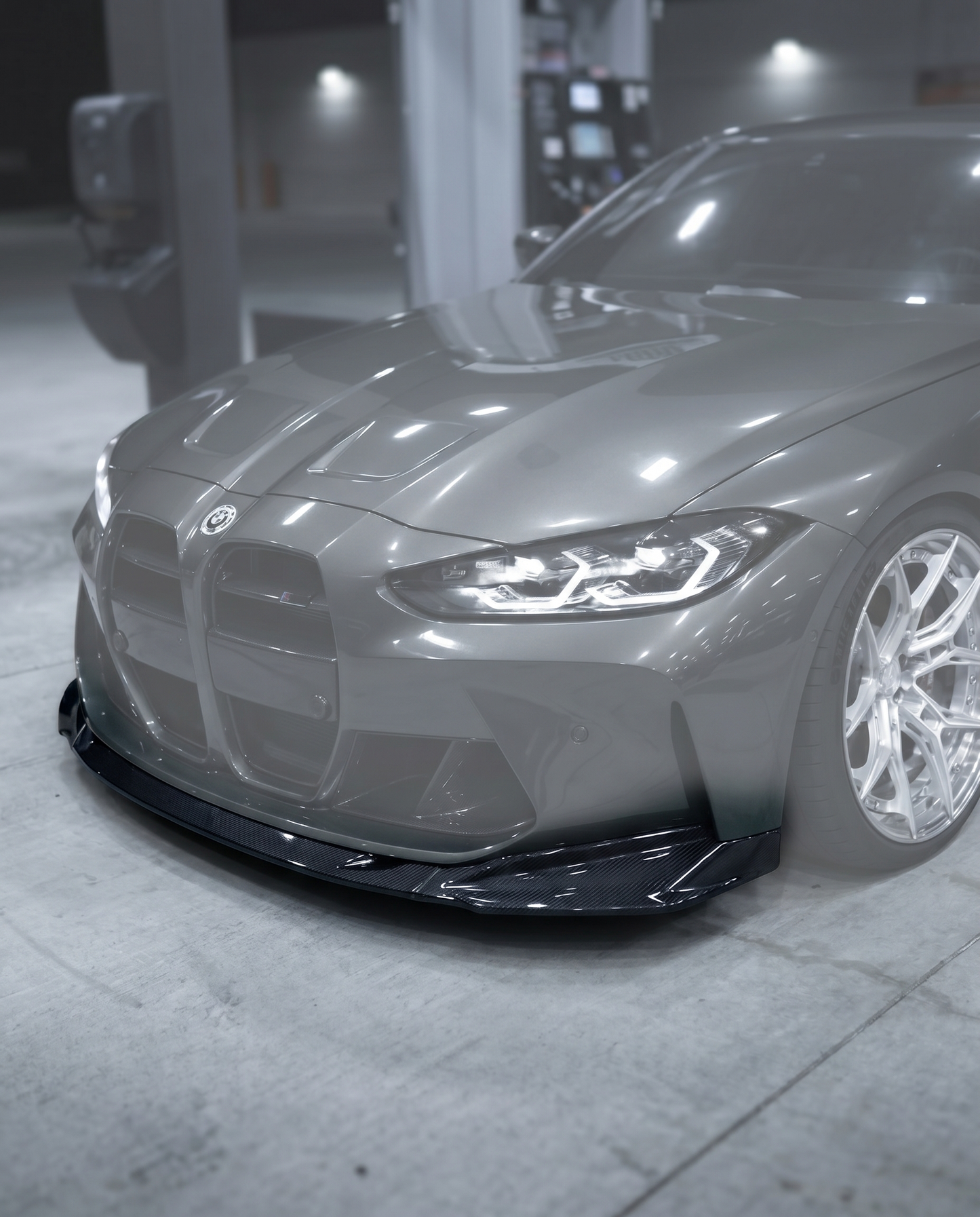 Gloss dry carbon front lip installed on BMW G80 M3 bumper, vehicle ghosted in blurred premium garage, OEM+ fitment shown sharp