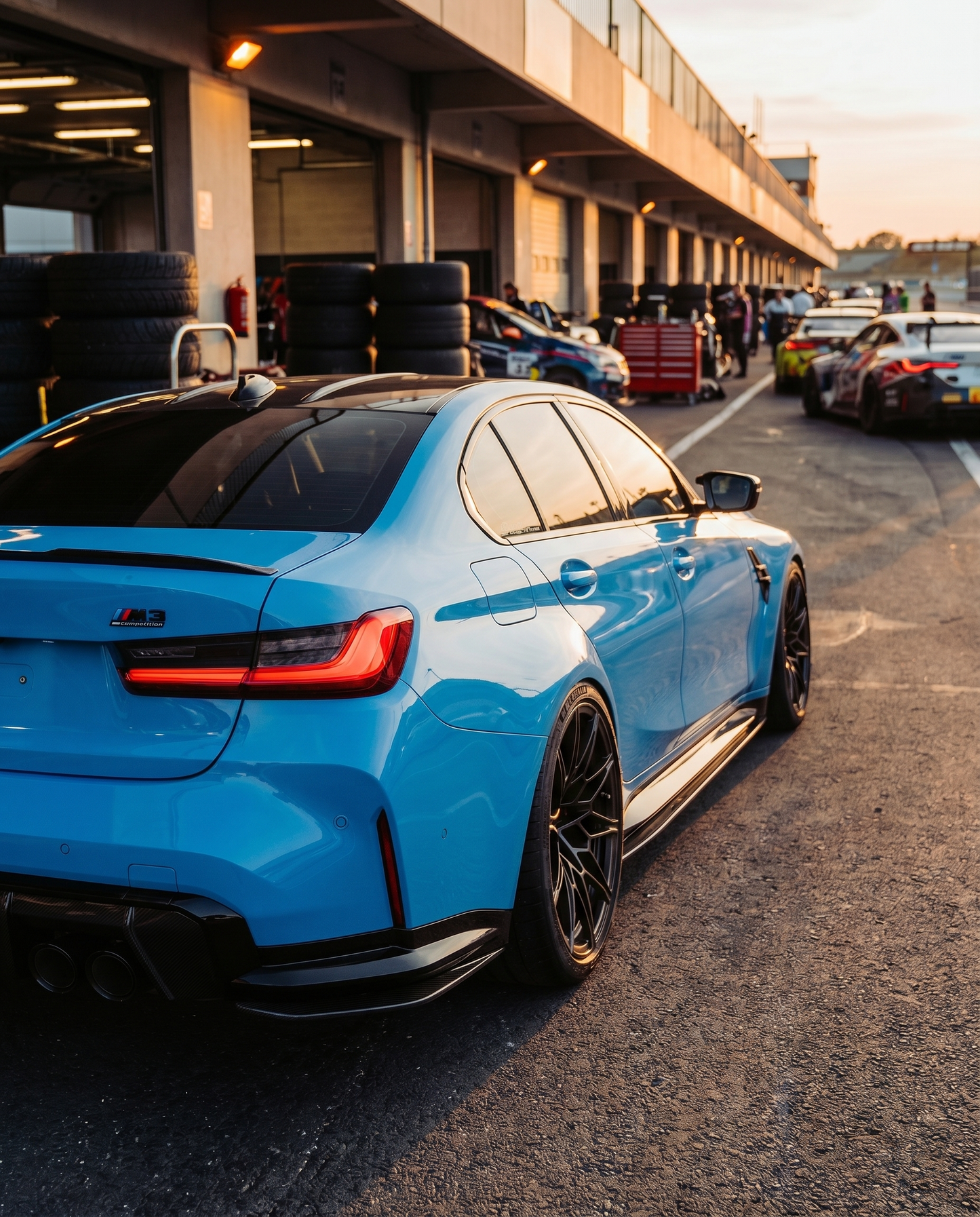 BMW G80 M3 rear 3/4 at dusk with gloss carbon diffuser side extensions, low raking light, aggressive motorsport rear-end stance