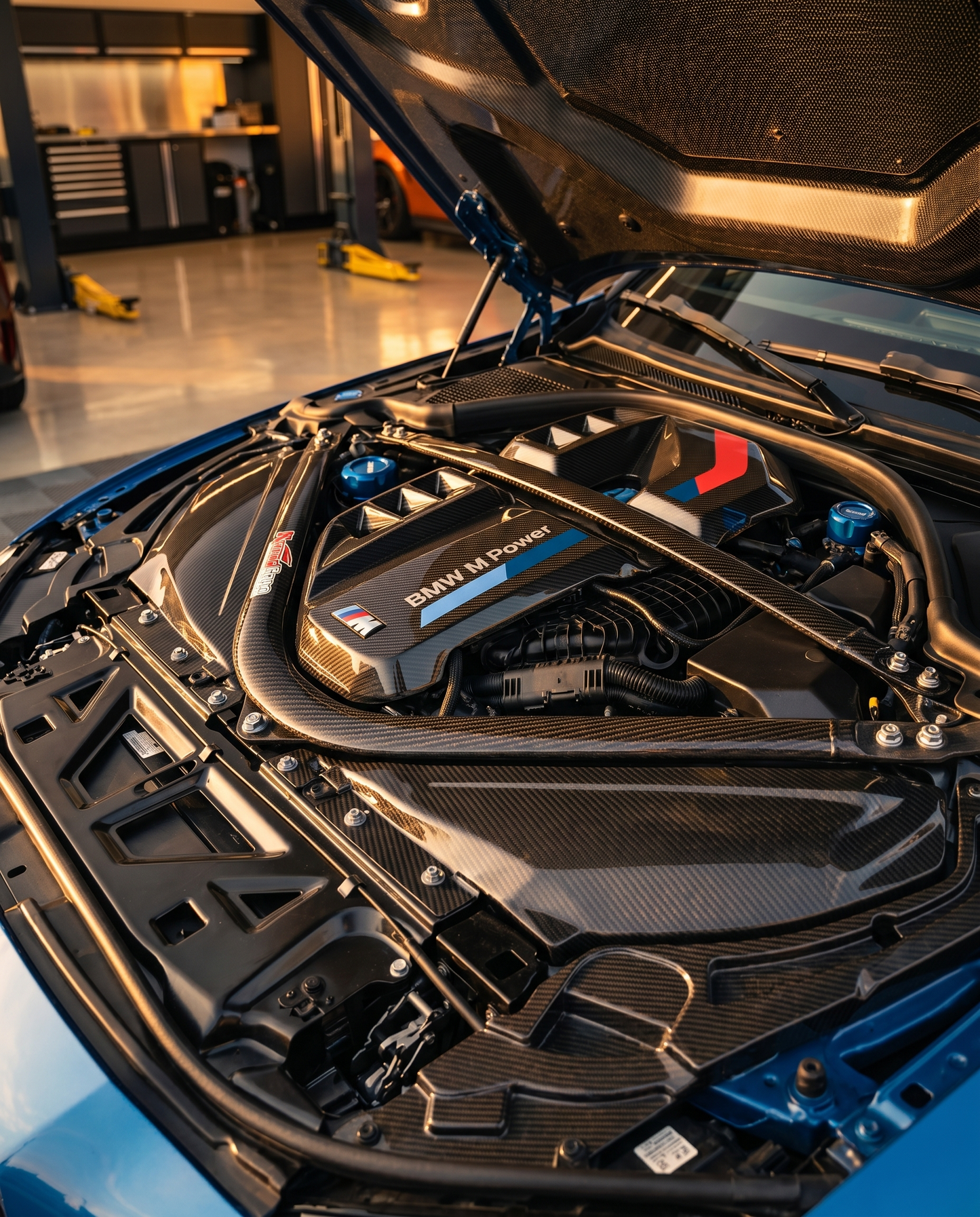 BMW G80 M3 engine bay at dusk with hood open showing gloss carbon air intake cover, warm raking light across the carbon weave