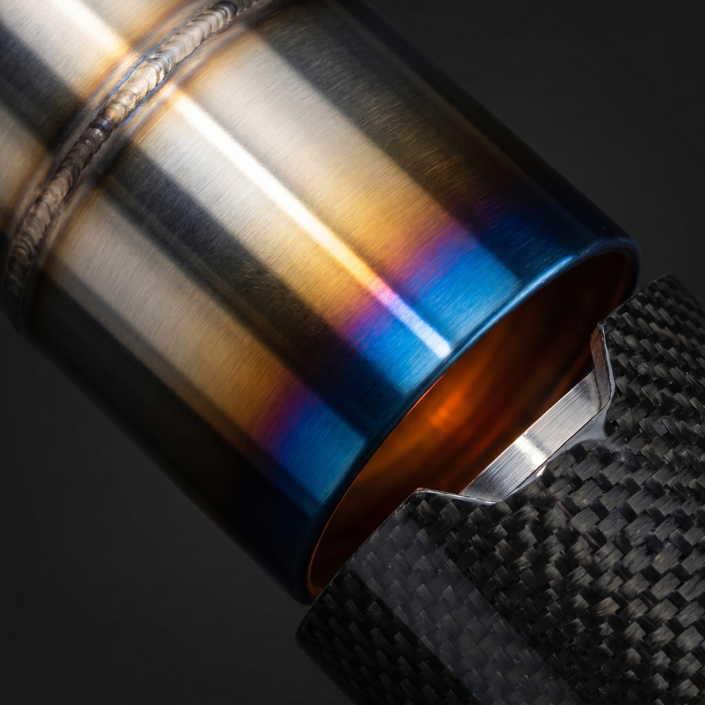 Macro of burnt titanium exhaust tip meeting gloss carbon sleeve on BMW G8X, blue-gold-silver heat gradient and precise clamp seam