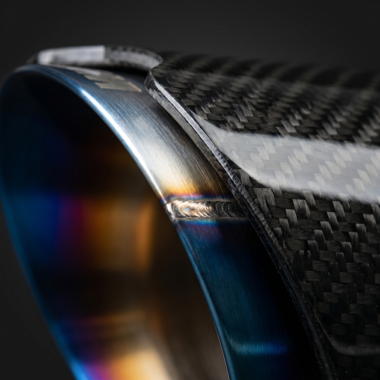 Macro of angle-cut burnt titanium exhaust tip meeting gloss carbon sleeve on BMW G8X, blue-gold-silver heat gradient and clamp seam