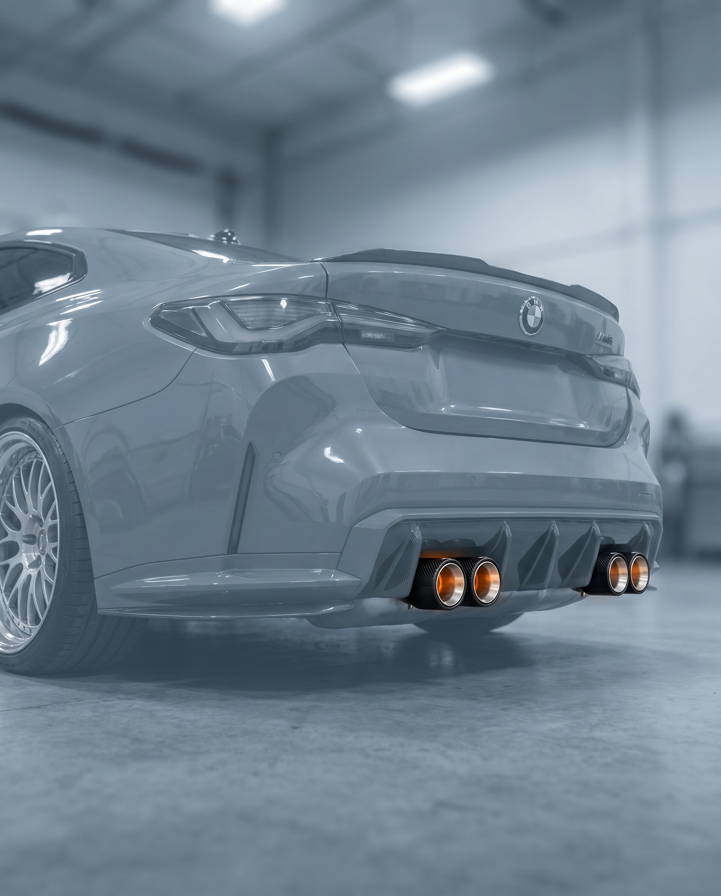 Oversized brushed aluminum carbon exhaust tips clamped on ghosted BMW G80 M3 rear, wide presence, blurred garage, OEM clamp-on fit