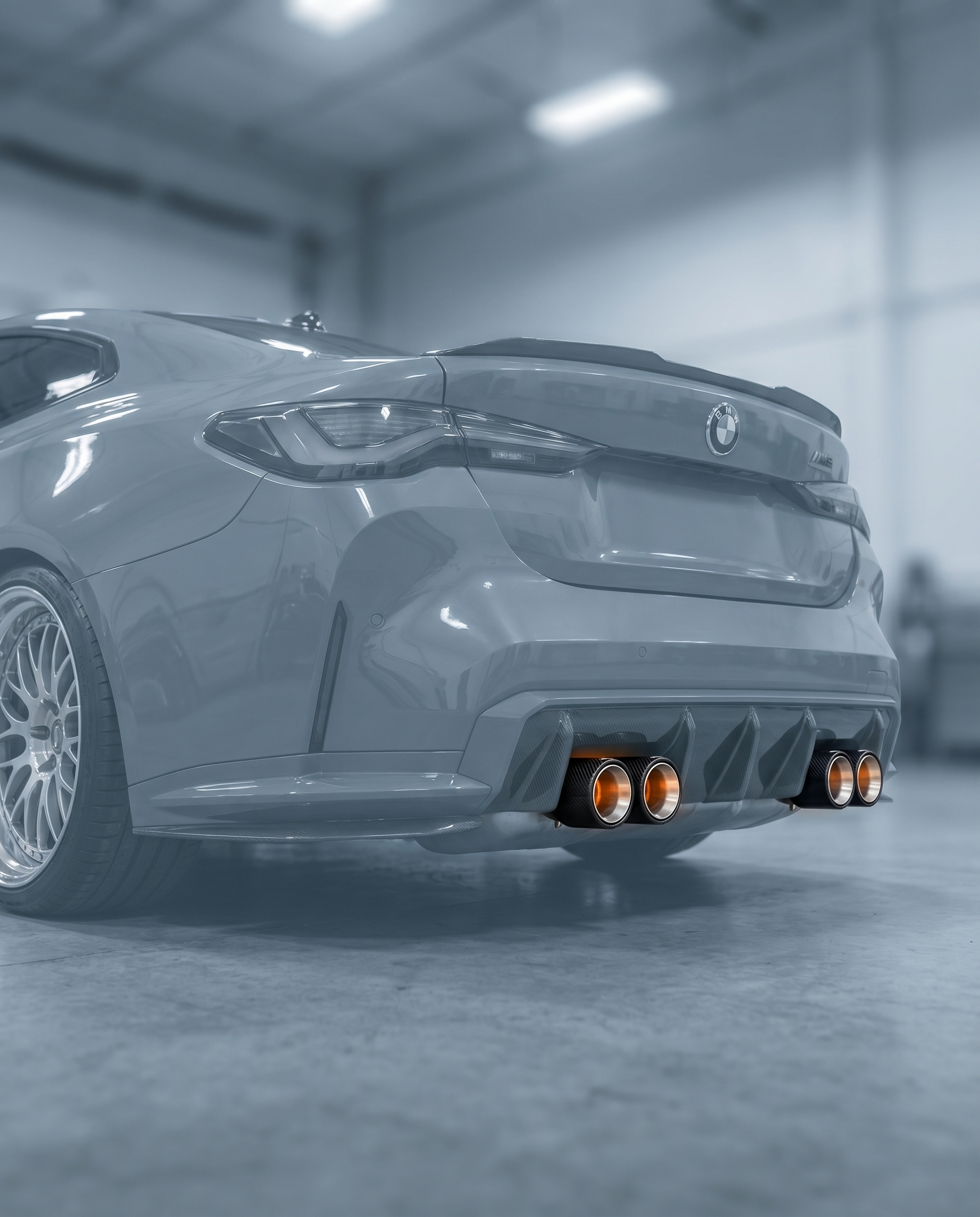 Oversized brushed aluminum carbon exhaust tips clamped on ghosted BMW G80 M3 rear, wide presence, blurred garage, OEM clamp-on fit