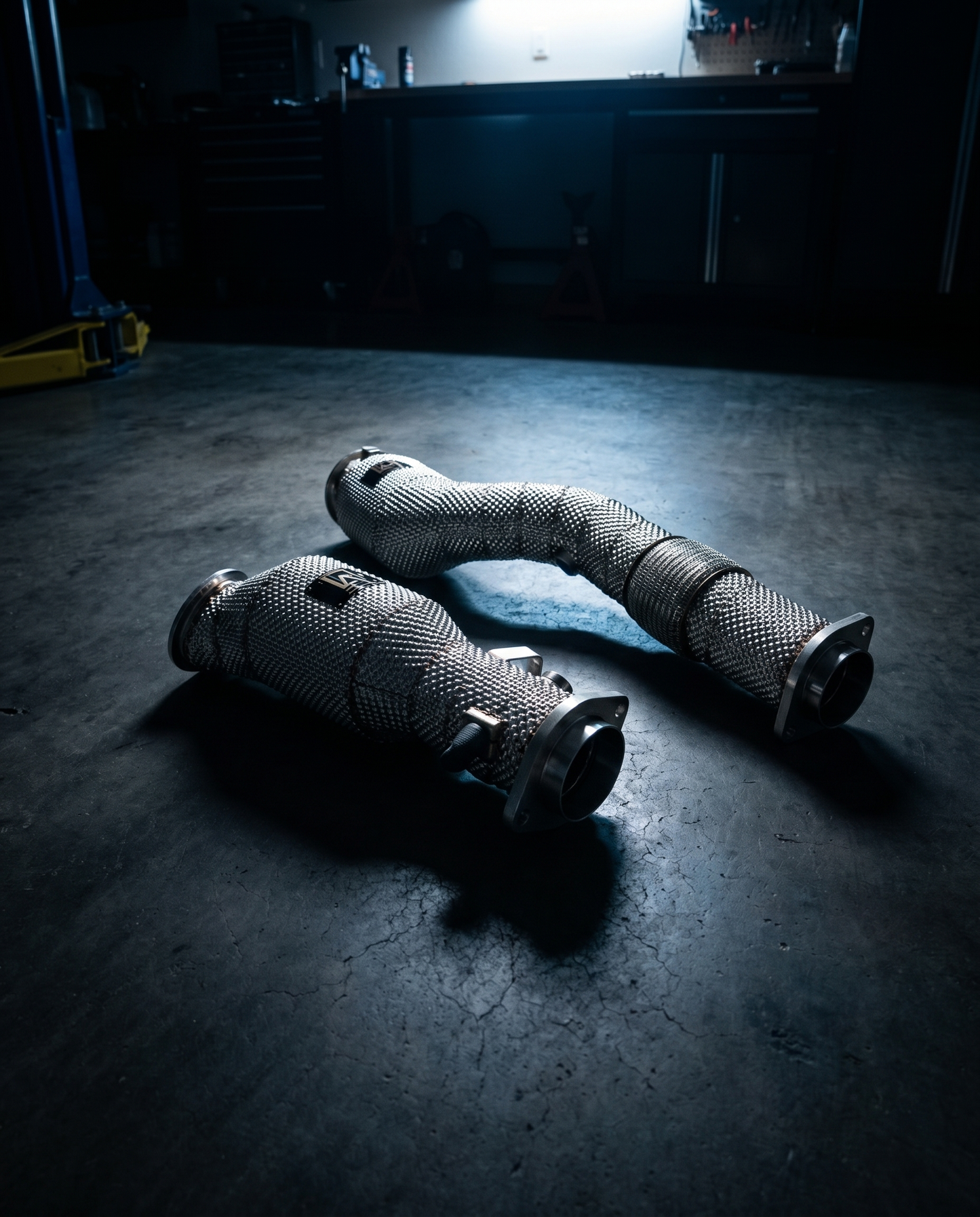 BMW S58 catted stainless downpipes on dark garage floor lit dramatically from above, mandrel bends and cat housings, precision part