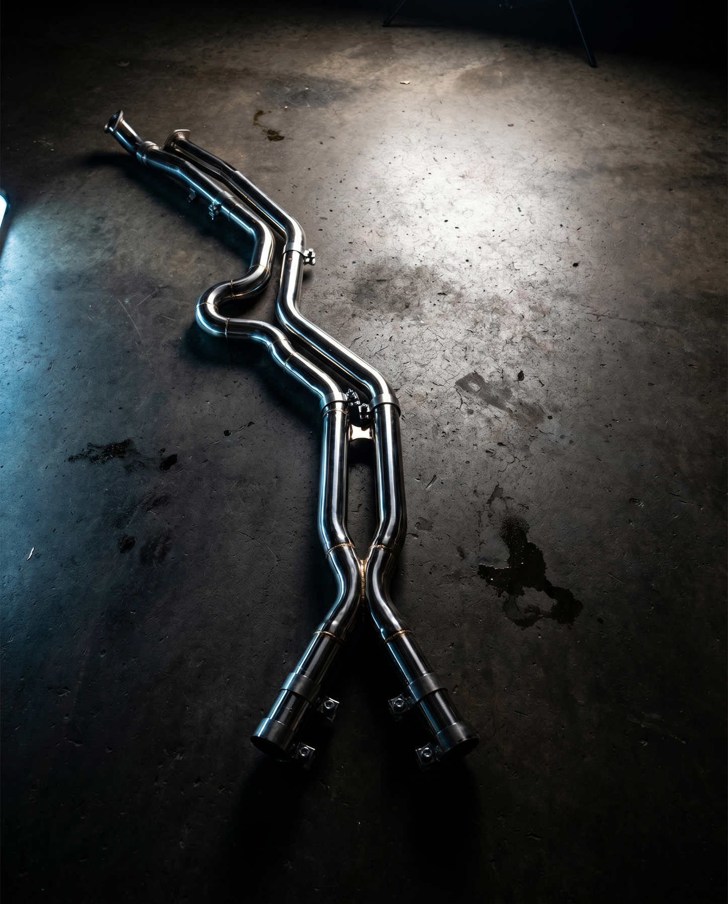 BMW G8X equal-length stainless midpipe on dark garage floor lit dramatically from above, polished bends and brace, precision part