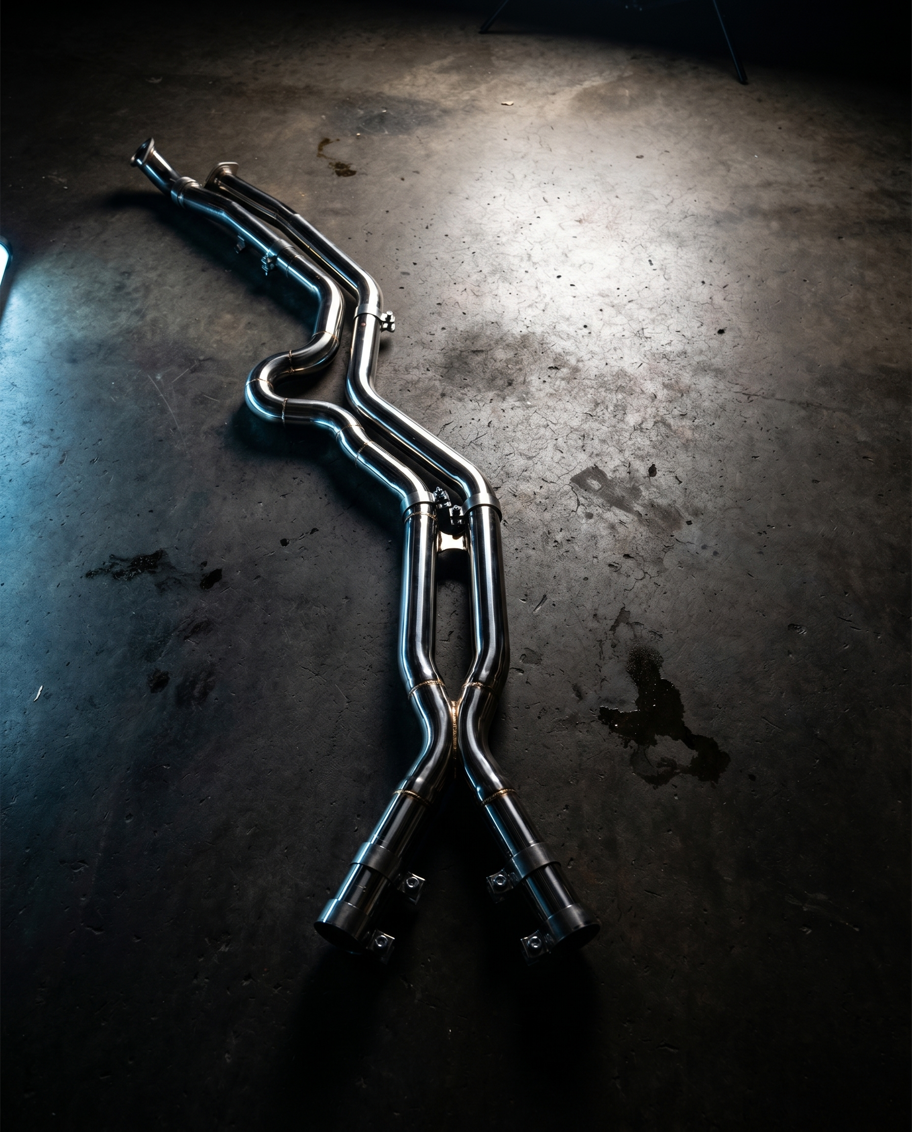 BMW G8X equal-length stainless midpipe on dark garage floor lit dramatically from above, polished bends and brace, precision part