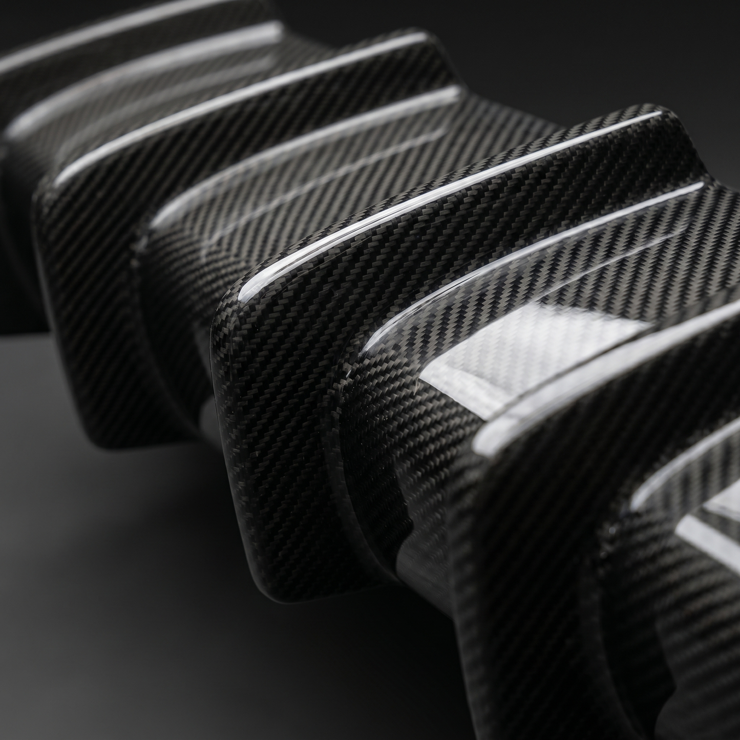 Macro of gloss 2x2 twill carbon weave over BMW M3 3D layered diffuser fins, clean light-bar reflection and machined fin edge
