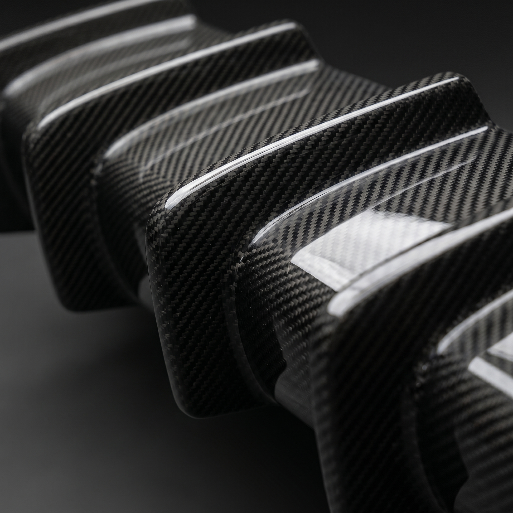 Macro of gloss 2x2 twill carbon weave over BMW M3 3D layered diffuser fins, clean light-bar reflection and machined fin edge
