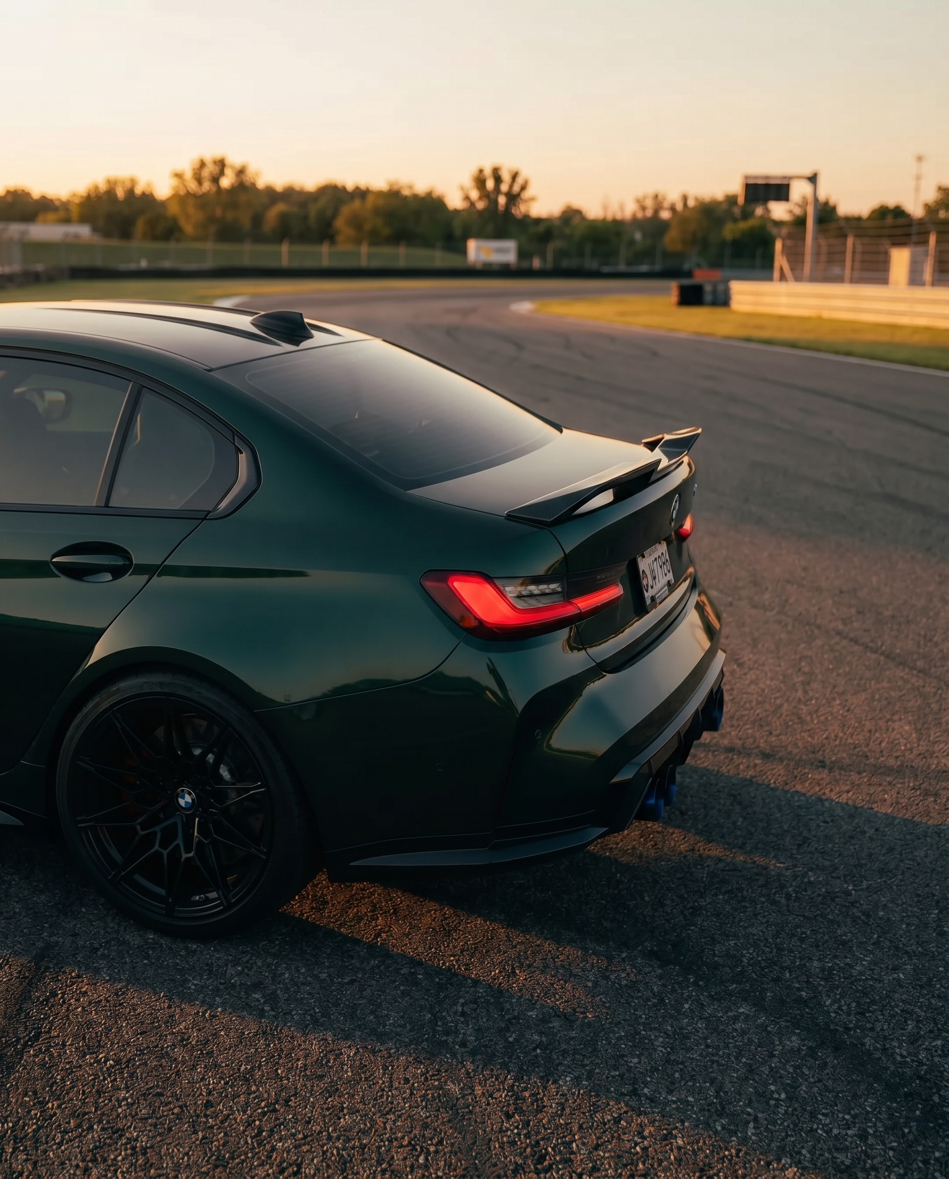 BMW G80 M3 at golden-hour dusk with gloss V-style carbon trunk spoiler, rear 3/4 view sharpening the silhouette